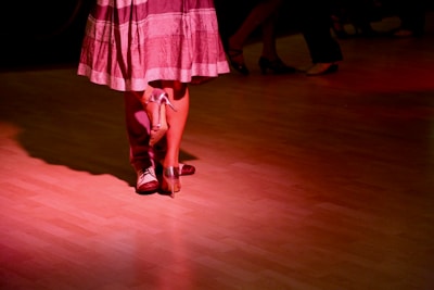 Two individuals are dancing closely on a polished wooden floor, illuminated by soft red lighting. The focus is on their feet; one wearing formal shoes and the other in high heels, suggesting a dance like the tango or waltz.