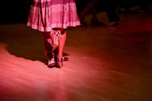 Two individuals are dancing closely on a polished wooden floor, illuminated by soft red lighting. The focus is on their feet; one wearing formal shoes and the other in high heels, suggesting a dance like the tango or waltz.