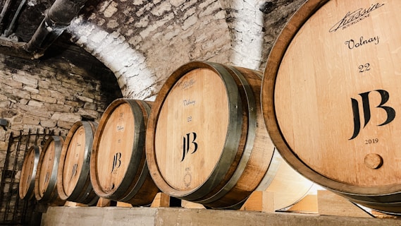 Wooden wine barrels are lined up against a stone wall in a dimly lit cellar. The barrels have logos and inscriptions on them, indicating a winery setting.