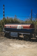 A tanker trailer is parked on a patch of dirt with wild grass and scrub in the background. The side of the tank is painted red and blue, with graffiti saying 'BURN PATRIARCHY' and an anarchy symbol. In the background, there is a vertical industrial chimney and utility poles under a clear blue sky.
