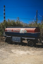 A tanker trailer is parked on a patch of dirt with wild grass and scrub in the background. The side of the tank is painted red and blue, with graffiti saying 'BURN PATRIARCHY' and an anarchy symbol. In the background, there is a vertical industrial chimney and utility poles under a clear blue sky.
