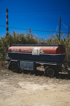 A tanker trailer is parked on a patch of dirt with wild grass and scrub in the background. The side of the tank is painted red and blue, with graffiti saying 'BURN PATRIARCHY' and an anarchy symbol. In the background, there is a vertical industrial chimney and utility poles under a clear blue sky.