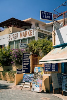 A street corner with a supermarket and a cafe bar. There are signs advertising various services and products, including ceramics, souvenirs, and local wines. Posters and boards display images and information for tourists, set against a backdrop of lush greenery and clear blue skies.