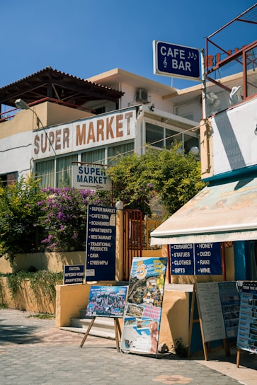 A street corner with a supermarket and a cafe bar. There are signs advertising various services and products, including ceramics, souvenirs, and local wines. Posters and boards display images and information for tourists, set against a backdrop of lush greenery and clear blue skies.