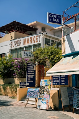 A street corner with a supermarket and a cafe bar. There are signs advertising various services and products, including ceramics, souvenirs, and local wines. Posters and boards display images and information for tourists, set against a backdrop of lush greenery and clear blue skies.