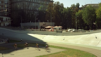 A group of cyclists is riding on an outdoor velodrome track. The scene is set on a sunny day, with shadows cast on the track. In the background, there is a modern building with a terrace, alongside parked cars and trees providing a lush green backdrop.