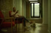 A person is sitting in a red chair holding a phone, with a small white dog lying on the floor beside them. The setting appears to be an indoor space with brick and white walls, wooden floors, and a partially visible green plant near a glass door.