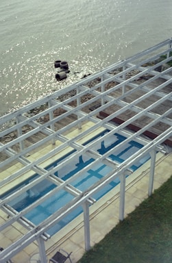 A body of water with shimmering reflections is bordered by a rocky shoreline. In the foreground, an empty swimming pool is visible beneath a wooden pergola structure. The area around the pool is paved, and a patch of green grass is seen to the right. Several tires float near the shoreline.