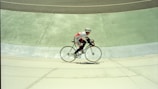 A cyclist in a racing outfit rides a bicycle on a sloped velodrome track. The motion blur indicates speed, and the background shows distinct sections of the track in varying shades of green and grey.
