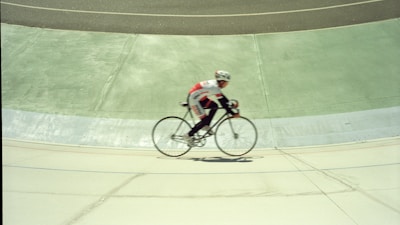 A cyclist in a racing outfit rides a bicycle on a sloped velodrome track. The motion blur indicates speed, and the background shows distinct sections of the track in varying shades of green and grey.
