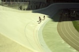 Bicyclists ride on a curved velodrome track under bright sunlight. The track is concrete with colored lines marking lanes. Spectators can be seen in the stands above, observing the event. Shadows create a distinct contrast on the left side of the image.