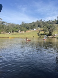 A serene lake scene with people engaging in recreational activities. A person is kayaking in the middle of the lake, while others engage in various outdoor activities on the grassy area surrounding the lake. The background features lush greenery and trees under a bright, clear sky.