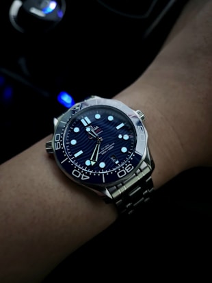 A wrist adorned with a luxury dive watch, featuring a dark blue dial with luminescent hour markers and hands. The watch is encased in polished stainless steel, and it includes a rotating bezel with prominent minute indicators. The background is dimly lit, with a hint of blue light from what appears to be a car's dashboard or control panel.