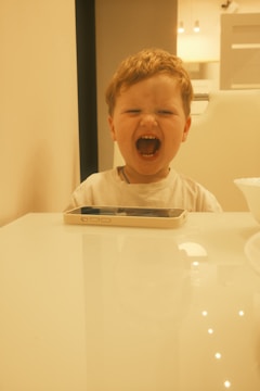 A young child with light brown hair is sitting behind a glossy table, their mouth open wide as if yelling or laughing. A smartphone is placed on the table. The background features soft lighting, contributing to a warm and cozy atmosphere.