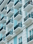 A modern apartment building featuring multiple balconies with black railings. The structure has a sleek, minimalist design using a combination of light blue and white colors, creating a clean, contemporary appearance.