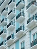 A modern apartment building featuring multiple balconies with black railings. The structure has a sleek, minimalist design using a combination of light blue and white colors, creating a clean, contemporary appearance.