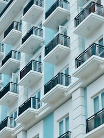 A modern apartment building featuring multiple balconies with black railings. The structure has a sleek, minimalist design using a combination of light blue and white colors, creating a clean, contemporary appearance.