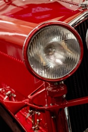 A close-up view of a classic car's headlight, with a rich red paint finish and polished chrome accents. The intricate design of the headlight features a ribbed glass cover, enhancing the vintage appeal. Reflections and highlights accentuate the glossy surface.
