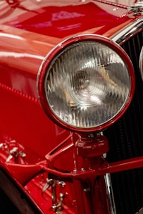 A close-up view of a classic car's headlight, with a rich red paint finish and polished chrome accents. The intricate design of the headlight features a ribbed glass cover, enhancing the vintage appeal. Reflections and highlights accentuate the glossy surface.