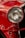 A close-up view of a classic car's headlight, with a rich red paint finish and polished chrome accents. The intricate design of the headlight features a ribbed glass cover, enhancing the vintage appeal. Reflections and highlights accentuate the glossy surface.