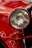 A close-up view of a classic car's headlight, with a rich red paint finish and polished chrome accents. The intricate design of the headlight features a ribbed glass cover, enhancing the vintage appeal. Reflections and highlights accentuate the glossy surface.