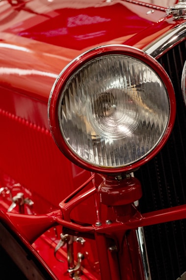 A close-up view of a classic car's headlight, with a rich red paint finish and polished chrome accents. The intricate design of the headlight features a ribbed glass cover, enhancing the vintage appeal. Reflections and highlights accentuate the glossy surface.