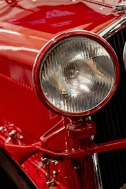 A close-up view of a classic car's headlight, with a rich red paint finish and polished chrome accents. The intricate design of the headlight features a ribbed glass cover, enhancing the vintage appeal. Reflections and highlights accentuate the glossy surface.
