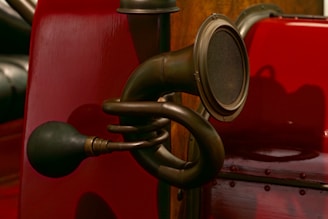 An antique-style horn with a rubber bulb attached to a brass tube, mounted on a vintage vehicle featuring a glossy red surface. The horn's intricate tubing is visible, and there's a rich wood texture in the background, suggesting an element of craftsmanship.