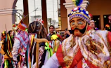 A colorful traditional celebration with people wearing ornate costumes and masks. The central figure wears a bright costume with intricate designs and a mask featuring a mustached face. Vibrant ribbons and feathers adorn their outfits, and they are surrounded by a festive crowd.