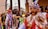 A colorful traditional celebration with people wearing ornate costumes and masks. The central figure wears a bright costume with intricate designs and a mask featuring a mustached face. Vibrant ribbons and feathers adorn their outfits, and they are surrounded by a festive crowd.