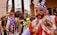A colorful traditional celebration with people wearing ornate costumes and masks. The central figure wears a bright costume with intricate designs and a mask featuring a mustached face. Vibrant ribbons and feathers adorn their outfits, and they are surrounded by a festive crowd.