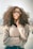 A fashionable woman with curly hair wearing stylish sunglasses and a beige sweater, striking a pose with one hand near her chin. The soft focus background adds a dreamy effect, emphasizing her confident and serene expression.