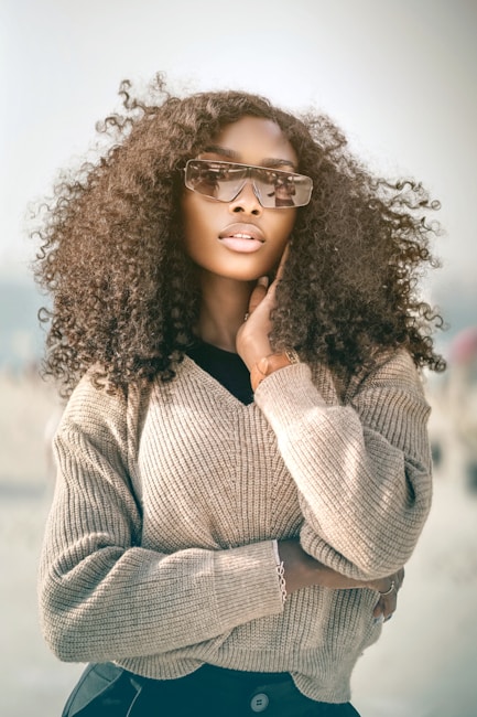 A fashionable woman with curly hair wearing stylish sunglasses and a beige sweater, striking a pose with one hand near her chin. The soft focus background adds a dreamy effect, emphasizing her confident and serene expression.