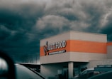 A supermarket building with a sign reading 'Multifood Supermarket' on its facade. The building features an orange accent strip and is positioned against a backdrop of dark, stormy clouds. Cars and power lines are visible in the foreground and background, adding to an urban setting.