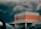 A supermarket building with a sign reading 'Multifood Supermarket' on its facade. The building features an orange accent strip and is positioned against a backdrop of dark, stormy clouds. Cars and power lines are visible in the foreground and background, adding to an urban setting.