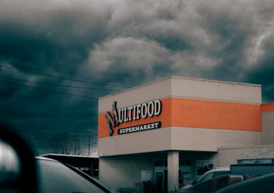 A supermarket building with a sign reading 'Multifood Supermarket' on its facade. The building features an orange accent strip and is positioned against a backdrop of dark, stormy clouds. Cars and power lines are visible in the foreground and background, adding to an urban setting.
