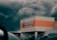 A supermarket building with a sign reading 'Multifood Supermarket' on its facade. The building features an orange accent strip and is positioned against a backdrop of dark, stormy clouds. Cars and power lines are visible in the foreground and background, adding to an urban setting.