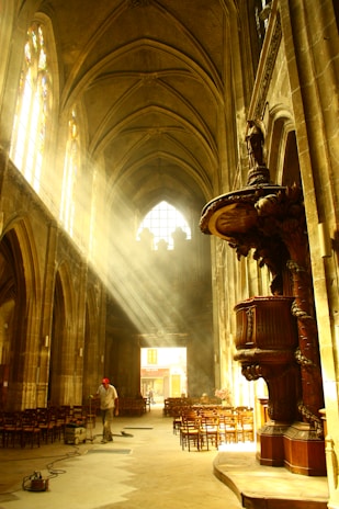 Sunlight streams through the stained glass windows of a cathedral, illuminating dust in the air and casting a warm glow on the interior. The vaulted ceilings and stone columns create a grand and reverent atmosphere. A person wearing a red cap is seen cleaning the floor amidst rows of wooden chairs.