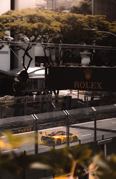 A bright yellow sports car speeding down a racetrack, surrounded by tall metal fences. Above, a large sign with the Rolex logo is visible. Trees with lush green foliage provide a backdrop, and the atmosphere is lively and dynamic, suggesting a high-energy race event.