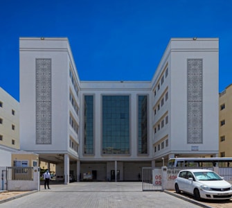 A large, symmetrical building with a modern architectural design, featuring a central glass facade flanked by two white sections with intricate decorative panels. The building is surrounded by a paved area with a car and a bus parked in front. A person stands near the entrance beside a security barrier.