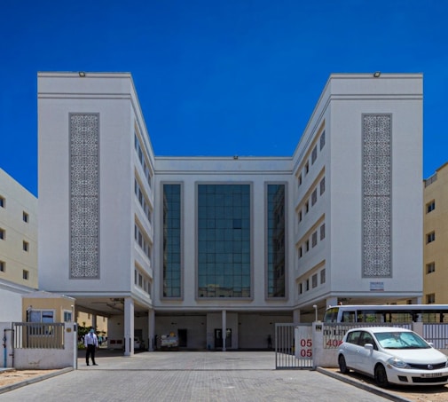 A large, symmetrical building with a modern architectural design, featuring a central glass facade flanked by two white sections with intricate decorative panels. The building is surrounded by a paved area with a car and a bus parked in front. A person stands near the entrance beside a security barrier.