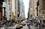 A busy urban street scene featuring numerous vehicles and pedestrians. High-rise buildings line both sides of the street, showcasing a mix of architectural styles. Traffic signals are visible, and people are engaged in various activities, contributing to the liveliness of the city environment.