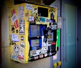 A heavily vandalized ticket machine covered in various colorful stickers and graffiti is mounted on a wall. The machine appears worn and features a keypad, display screen, and card slot. Surrounding the machine, there are smudges and dirt accumulated over time, contributing to its neglected appearance.