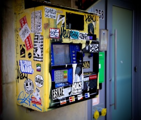 A heavily vandalized ticket machine covered in various colorful stickers and graffiti is mounted on a wall. The machine appears worn and features a keypad, display screen, and card slot. Surrounding the machine, there are smudges and dirt accumulated over time, contributing to its neglected appearance.