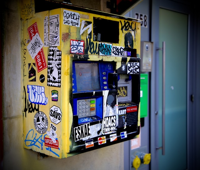 A heavily vandalized ticket machine covered in various colorful stickers and graffiti is mounted on a wall. The machine appears worn and features a keypad, display screen, and card slot. Surrounding the machine, there are smudges and dirt accumulated over time, contributing to its neglected appearance.
