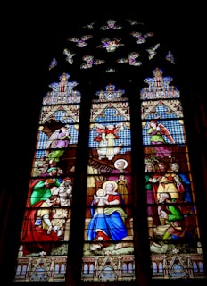 A stained glass window depicting a biblical scene with various characters dressed in traditional robes. At the center, a woman holding a child sits calmly, surrounded by several figures, some of whom appear to be kneeling or presenting gifts. The scene is intricately designed with ornate patterns and vibrant colors.