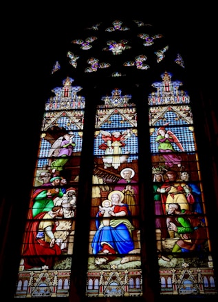 A stained glass window depicting a biblical scene with various characters dressed in traditional robes. At the center, a woman holding a child sits calmly, surrounded by several figures, some of whom appear to be kneeling or presenting gifts. The scene is intricately designed with ornate patterns and vibrant colors.