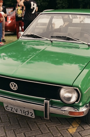 A vintage green Volkswagen car is parked on a brick-paved area with people standing in the background. The car's front grille prominently displays the VW emblem, and there is a license plate visible. The vehicle is in good condition, reflecting light slightly, and other cars and trees can be seen in the background.