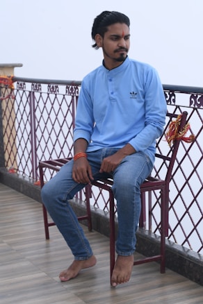 A man is sitting on a chair placed next to a decorative railing adorned with orange and yellow ribbons. He is wearing a light blue long-sleeve shirt with a logo, jeans, and an orange bracelet. His expression is contemplative while glancing to his right.