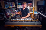 A person is sitting in a home studio surrounded by various musical instruments and equipment. The setup includes synthesizers, a mixing console, and a guitar. The person appears to be focusing on a synthesizer while smiling pleasantly, creating a creative and enthusiastic atmosphere.